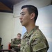 A U.S. Army soldier in Operational Camouflage Pattern (OCP) uniform is standing in what appears to be a classroom or briefing room. The soldier's name tape reads 'KOO' and displays a Staff Sergeant rank insignia along with a '2nd Infantry Division' shoulder sleeve insignia. A second soldier is visible seated in the background.