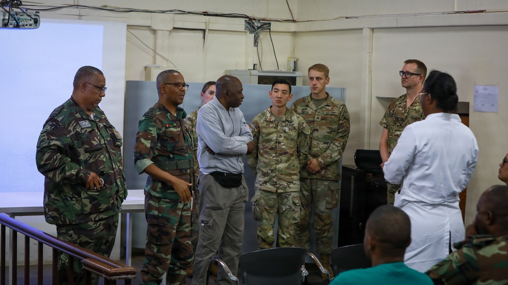A group of military personnel from at least two different armed forces are gathered in what appears to be a briefing or meeting room. The group includes soldiers in US Army MultiCam/OCP uniforms and others in a distinct camouflage pattern, along with a civilian and a person in a white coat. They appear to be engaged in a discussion or presentation, with a projection screen visible in the background.