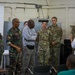 A group of military personnel from at least two different armed forces are gathered in what appears to be a briefing or meeting room. The group includes soldiers in US Army MultiCam/OCP uniforms and others in a distinct camouflage pattern, along with a civilian and a person in a white coat. They appear to be engaged in a discussion or presentation, with a projection screen visible in the background.
