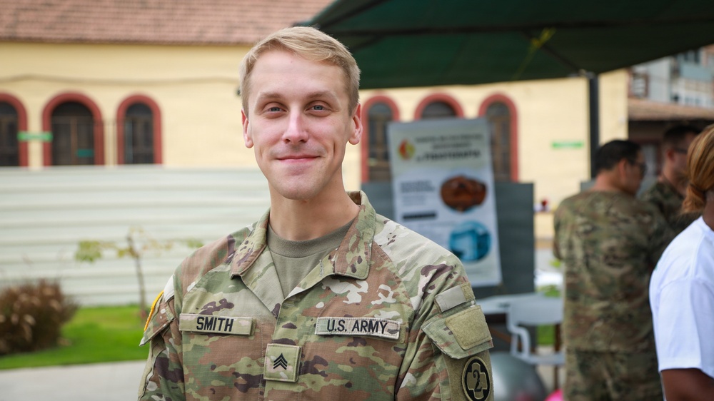 A U.S. Army soldier identified by name tape as 'SMITH' poses for a photo in an outdoor setting. He is wearing the Operational Camouflage Pattern (OCP) uniform and displays the rank of Sergeant on his chest. The background shows a European-style building and other military personnel.