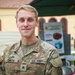A U.S. Army soldier identified by name tape as 'SMITH' poses for a photo in an outdoor setting. He is wearing the Operational Camouflage Pattern (OCP) uniform and displays the rank of Sergeant on his chest. The background shows a European-style building and other military personnel.