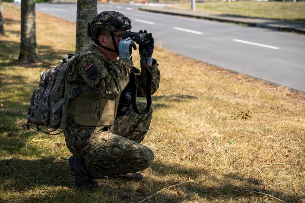 A soldier in full combat gear is crouching near a tree line alongside a road, using binoculars to observe a distant area. The soldier is equipped with a tactical vest, camouflage uniform, helmet, backpack, and gloves. A small flag patch is visible on the left shoulder of the uniform.