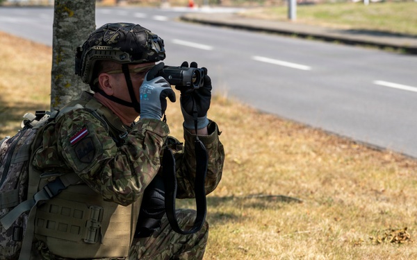 A soldier in full combat gear is crouching near a tree line alongside a road, using binoculars to observe a distant area. The soldier is equipped with a tactical vest, camouflage uniform, helmet, backpack, and gloves. A small flag patch is visible on the left shoulder of the uniform.