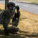 A soldier in full combat gear is crouching near a tree line alongside a road, using binoculars to observe a distant area. The soldier is equipped with a tactical vest, camouflage uniform, helmet, backpack, and gloves. A small flag patch is visible on the left shoulder of the uniform.