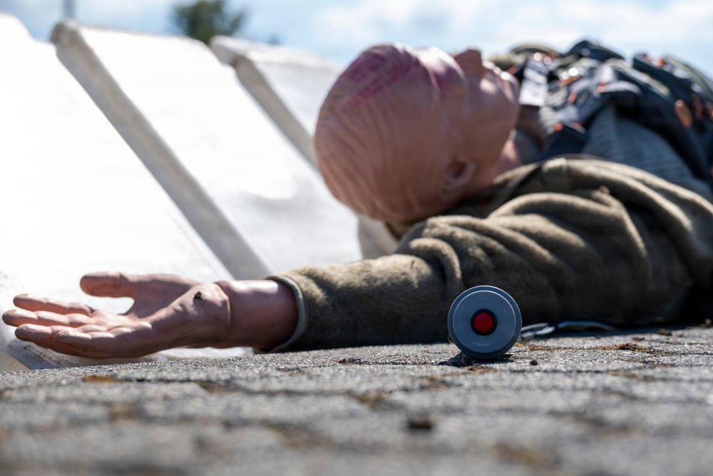 The image depicts a training or simulation scenario featuring a mannequin or dummy dressed in clothing, lying on a concrete surface outdoors. A small cylindrical device with a red button is visible near the outstretched hand of the figure, suggesting an emergency alert or medical alarm context. White panels or boards are visible in the background, consistent with a staged training exercise.