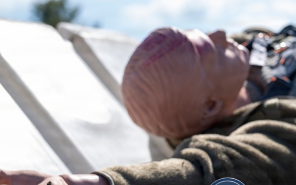 The image depicts a training or simulation scenario featuring a mannequin or dummy dressed in clothing, lying on a concrete surface outdoors. A small cylindrical device with a red button is visible near the outstretched hand of the figure, suggesting an emergency alert or medical alarm context. White panels or boards are visible in the background, consistent with a staged training exercise.