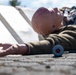 The image depicts a training or simulation scenario featuring a mannequin or dummy dressed in clothing, lying on a concrete surface outdoors. A small cylindrical device with a red button is visible near the outstretched hand of the figure, suggesting an emergency alert or medical alarm context. White panels or boards are visible in the background, consistent with a staged training exercise.