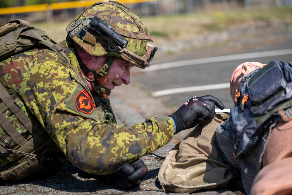A soldier in digital camouflage uniform is lying prone on a paved surface, appearing to administer first aid or conduct a close inspection of another person or object. The soldier is wearing full tactical gear including a helmet with mounted devices, black gloves, and a backpack. This appears to be a military training exercise or field medical scenario.