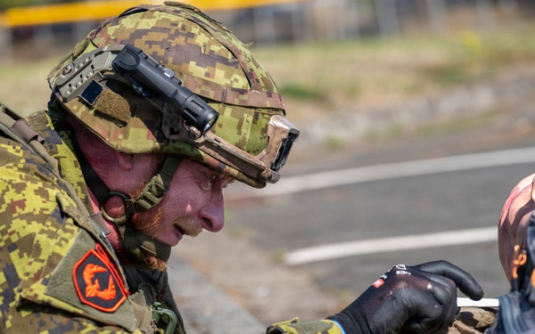 A soldier in digital camouflage uniform is lying prone on a paved surface, appearing to administer first aid or conduct a close inspection of another person or object. The soldier is wearing full tactical gear including a helmet with mounted devices, black gloves, and a backpack. This appears to be a military training exercise or field medical scenario.