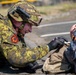 A soldier in digital camouflage uniform is lying prone on a paved surface, appearing to administer first aid or conduct a close inspection of another person or object. The soldier is wearing full tactical gear including a helmet with mounted devices, black gloves, and a backpack. This appears to be a military training exercise or field medical scenario.