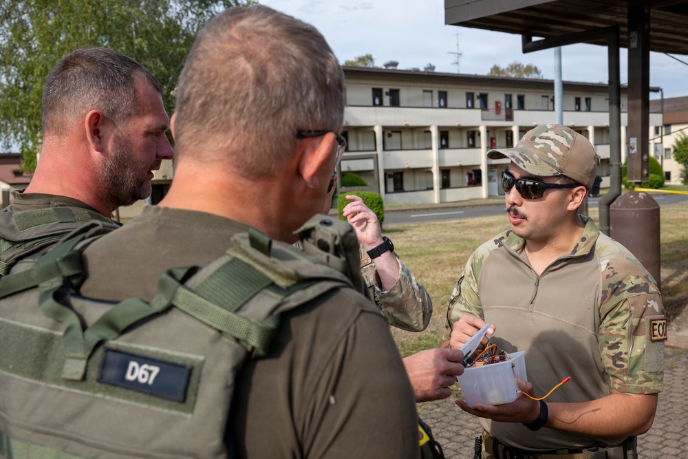 Three military personnel in tactical gear are engaged in a discussion outdoors near a multi-story building, likely on a military installation. One individual wearing a patch labeled 'EOD' (Explosive Ordnance Disposal) is holding a clear container with wires and components inside. The group appears to be conducting a briefing or training exercise related to EOD operations.