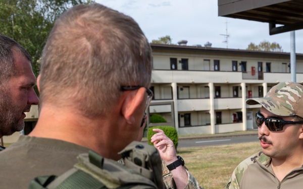 Three military personnel in tactical gear are engaged in a discussion outdoors near a multi-story building, likely on a military installation. One individual wearing a patch labeled 'EOD' (Explosive Ordnance Disposal) is holding a clear container with wires and components inside. The group appears to be conducting a briefing or training exercise related to EOD operations.