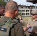 Three military personnel in tactical gear are engaged in a discussion outdoors near a multi-story building, likely on a military installation. One individual wearing a patch labeled 'EOD' (Explosive Ordnance Disposal) is holding a clear container with wires and components inside. The group appears to be conducting a briefing or training exercise related to EOD operations.