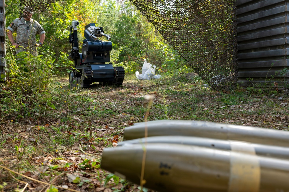 A military EOD (Explosive Ordnance Disposal) scenario showing a tracked robot being used to investigate or handle suspected ordnance. In the foreground, two cylindrical projectiles (likely artillery shells or mortar rounds) are visible on the ground. A soldier in camouflage uniform observes from a safe distance while the robot operates near white bags in the background.