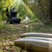 A military EOD (Explosive Ordnance Disposal) scenario showing a tracked robot being used to investigate or handle suspected ordnance. In the foreground, two cylindrical projectiles (likely artillery shells or mortar rounds) are visible on the ground. A soldier in camouflage uniform observes from a safe distance while the robot operates near white bags in the background.
