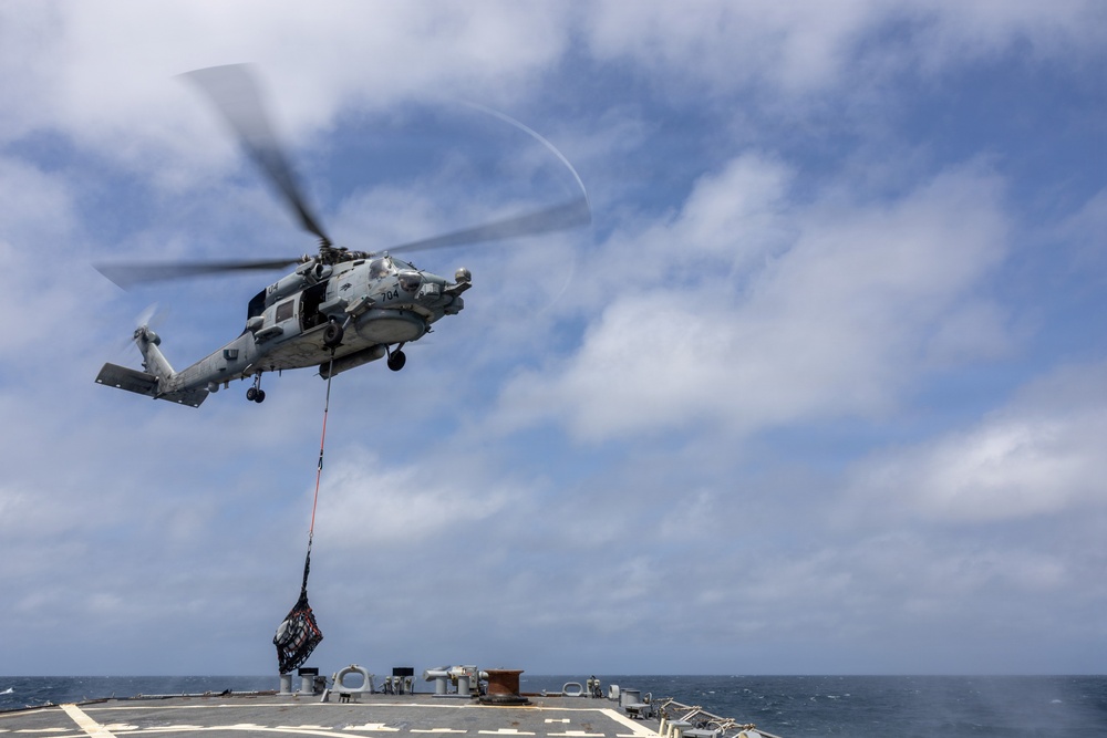 A military helicopter, marked with the number '704', is performing a vertical replenishment (VERTREP) operation over the deck of a naval vessel at sea. The helicopter is lowering a cargo net loaded with supplies onto the ship's deck using a hoist line. The scene takes place under partly cloudy skies over open ocean.