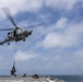 A military helicopter, marked with the number '704', is performing a vertical replenishment (VERTREP) operation over the deck of a naval vessel at sea. The helicopter is lowering a cargo net loaded with supplies onto the ship's deck using a hoist line. The scene takes place under partly cloudy skies over open ocean.