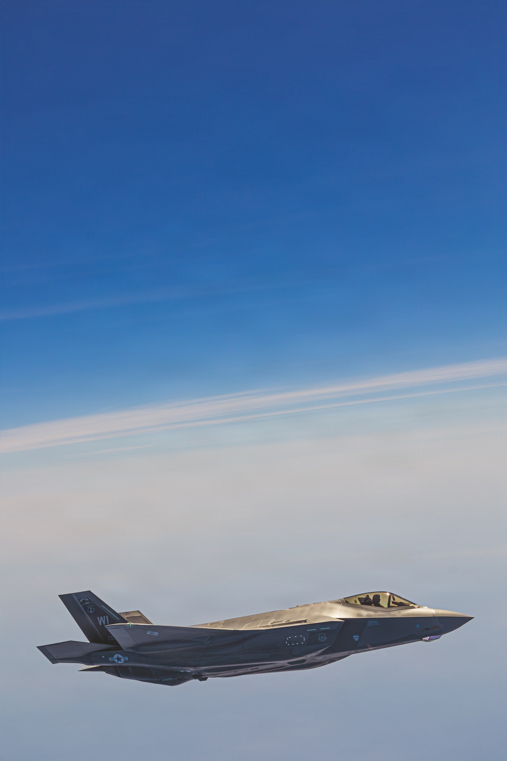A single F-35 Lightning II stealth fighter jet is photographed in flight against a clear blue sky with thin cirrus cloud layers below. The aircraft displays U.S. Air Force markings and tail code 'WI', indicating assignment to a Wisconsin Air National Guard unit. The image is a high-quality air-to-air photograph taken from close proximity, likely from another aircraft.