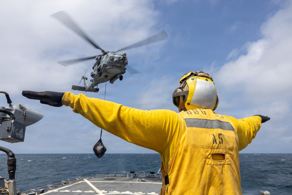 A naval deck crew member in a yellow flight deck jersey and helmet is directing a military helicopter during a vertical replenishment (VERTREP) operation at sea. The helicopter, appearing to be an MH-60 Seahawk, is hovering with a cargo net suspended beneath it. The operation is taking place on the deck of what appears to be a U.S. Navy destroyer or guided-missile destroyer.