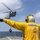 A naval deck crew member in a yellow flight deck jersey and helmet is directing a military helicopter during a vertical replenishment (VERTREP) operation at sea. The helicopter, appearing to be an MH-60 Seahawk, is hovering with a cargo net suspended beneath it. The operation is taking place on the deck of what appears to be a U.S. Navy destroyer or guided-missile destroyer.