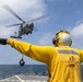 A naval deck crew member in a yellow flight deck jersey and helmet is directing a military helicopter during a vertical replenishment (VERTREP) operation at sea. The helicopter, appearing to be an MH-60 Seahawk, is hovering with a cargo net suspended beneath it. The operation is taking place on the deck of what appears to be a U.S. Navy destroyer or guided-missile destroyer.