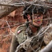 A soldier wearing Polish military uniform and tactical gear is positioned in a concealed observation post among dry branches and brush. The soldier appears alert and scanning the surroundings, with red rocky terrain visible in the background. The image was taken through tangled branches, creating a partially obscured perspective consistent with a field exercise or operational environment.