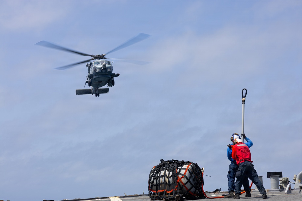 A military helicopter, appearing to be a naval variant such as an MH-60 Seahawk, hovers above a ship's flight deck during what appears to be a vertical replenishment (VERTREP) or cargo transfer operation. Two deck crew members in colored safety vests are visible on the deck, one holding a static discharge pole upward toward the helicopter. A netted cargo bundle secured with straps sits on the deck nearby.