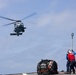 A military helicopter, appearing to be a naval variant such as an MH-60 Seahawk, hovers above a ship's flight deck during what appears to be a vertical replenishment (VERTREP) or cargo transfer operation. Two deck crew members in colored safety vests are visible on the deck, one holding a static discharge pole upward toward the helicopter. A netted cargo bundle secured with straps sits on the deck nearby.
