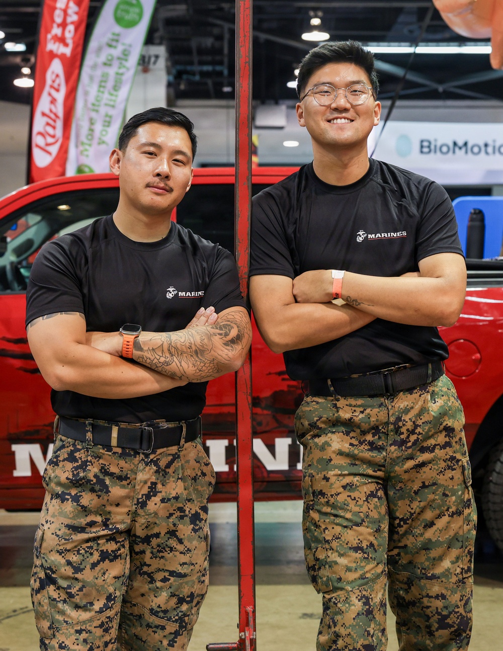 Two individuals wearing U.S. Marines branded black t-shirts and MARPAT (Marine Pattern) digital camouflage trousers stand with arms crossed in front of a red Marines-branded vehicle at what appears to be an indoor expo or recruitment event. Both are in a relaxed, confident pose leaning against a red metal pole. The setting includes visible sponsor banners including Ralphs and BioMotion in the background.