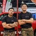 Two individuals wearing U.S. Marines branded black t-shirts and MARPAT (Marine Pattern) digital camouflage trousers stand with arms crossed in front of a red Marines-branded vehicle at what appears to be an indoor expo or recruitment event. Both are in a relaxed, confident pose leaning against a red metal pole. The setting includes visible sponsor banners including Ralphs and BioMotion in the background.