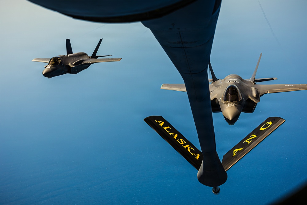 An aerial refueling operation is depicted, showing two F-35 Lightning II fighter jets flying in formation with a tanker aircraft. The tanker's refueling boom is visible in the foreground, marked with 'ALASKA' and 'ANG' (Alaska Air National Guard) in yellow lettering. One F-35 is positioned close to the boom for refueling while the second flies in escort formation to the left.