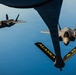 An aerial refueling operation is depicted, showing two F-35 Lightning II fighter jets flying in formation with a tanker aircraft. The tanker's refueling boom is visible in the foreground, marked with 'ALASKA' and 'ANG' (Alaska Air National Guard) in yellow lettering. One F-35 is positioned close to the boom for refueling while the second flies in escort formation to the left.