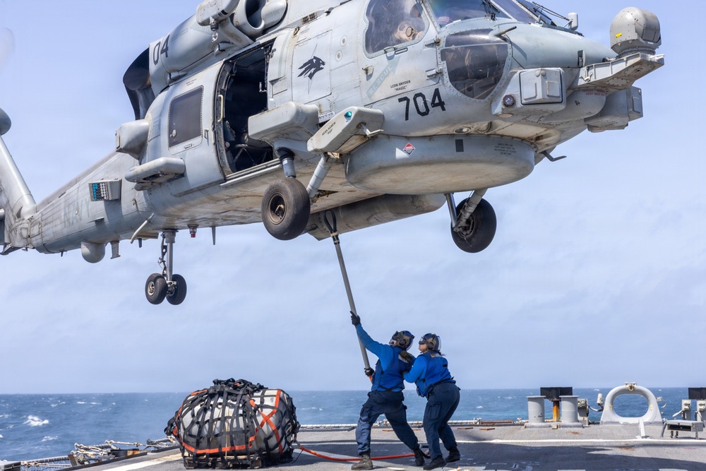 A U.S. Navy helicopter, numbered 704, hovers low over the deck of a naval vessel while two deck crew members in blue uniforms use a long pole to guide or stabilize the aircraft. A cargo net bundle secured with orange straps sits on the deck nearby, suggesting a vertical replenishment (VERTREP) or cargo transfer operation. The scene takes place at sea under a hazy sky with visible ocean in the background.