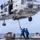 A U.S. Navy helicopter, numbered 704, hovers low over the deck of a naval vessel while two deck crew members in blue uniforms use a long pole to guide or stabilize the aircraft. A cargo net bundle secured with orange straps sits on the deck nearby, suggesting a vertical replenishment (VERTREP) or cargo transfer operation. The scene takes place at sea under a hazy sky with visible ocean in the background.