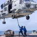 A U.S. Navy helicopter, numbered 704, hovers low over the deck of a naval vessel while two deck crew members in blue uniforms use a long pole to guide or stabilize the aircraft. A cargo net bundle secured with orange straps sits on the deck nearby, suggesting a vertical replenishment (VERTREP) or cargo transfer operation. The scene takes place at sea under a hazy sky with visible ocean in the background.