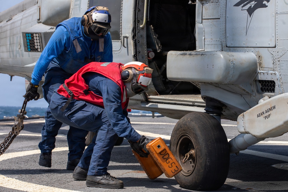 Two naval personnel are working on the landing gear of a military helicopter on a ship's flight deck. One sailor in a blue jumpsuit holds a chain while another in a red vest places or removes a wheel chock marked 'DDG53' from the helicopter's tire. The scene takes place aboard USS John Paul Jones (DDG-53), identifiable from the chock markings.