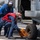 Two naval personnel are working on the landing gear of a military helicopter on a ship's flight deck. One sailor in a blue jumpsuit holds a chain while another in a red vest places or removes a wheel chock marked 'DDG53' from the helicopter's tire. The scene takes place aboard USS John Paul Jones (DDG-53), identifiable from the chock markings.