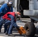 Two naval personnel are working on the landing gear of a military helicopter on a ship's flight deck. One sailor in a blue jumpsuit holds a chain while another in a red vest places or removes a wheel chock marked 'DDG53' from the helicopter's tire. The scene takes place aboard USS John Paul Jones (DDG-53), identifiable from the chock markings.