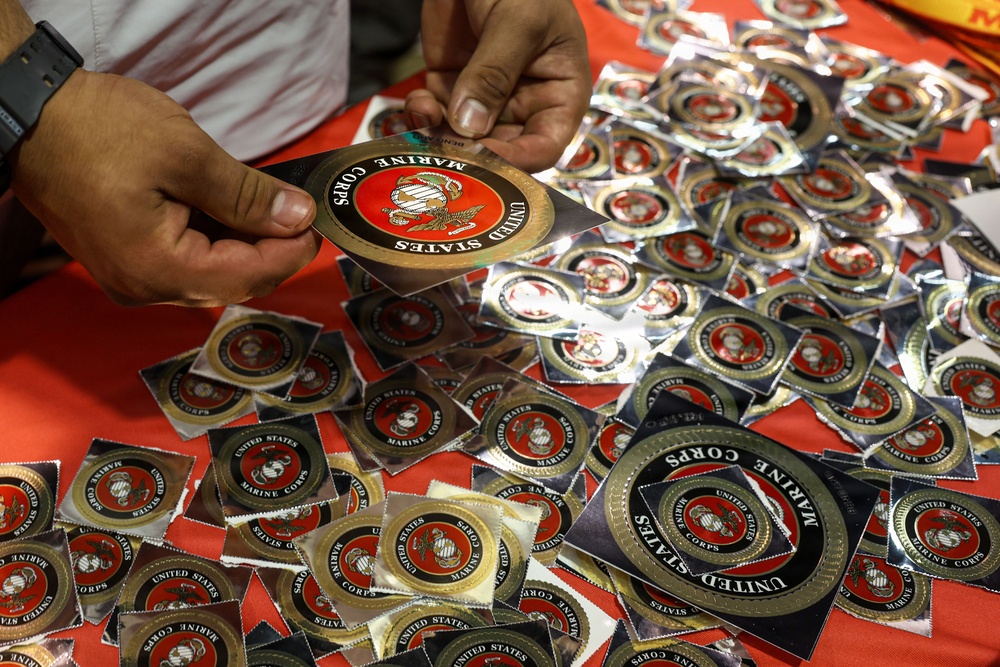 A person is handling United States Marine Corps emblem stickers/decals spread across a red surface. The stickers feature the official USMC Eagle, Globe, and Anchor emblem on a red and gold circular design. Multiple stickers are visible in individual clear plastic packaging, suggesting a distribution or display setting.