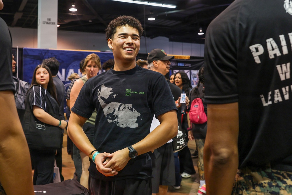 A young man wearing a black 'YEAT LOVE/LYFE TOUR 2026' t-shirt is smiling and appears to be engaged in conversation at what looks like an indoor convention or expo event. The setting is a large hall with multiple people milling about, booths or displays in the background, and overhead lighting. The atmosphere appears casual and social.