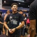A young man wearing a black 'YEAT LOVE/LYFE TOUR 2026' t-shirt is smiling and appears to be engaged in conversation at what looks like an indoor convention or expo event. The setting is a large hall with multiple people milling about, booths or displays in the background, and overhead lighting. The atmosphere appears casual and social.