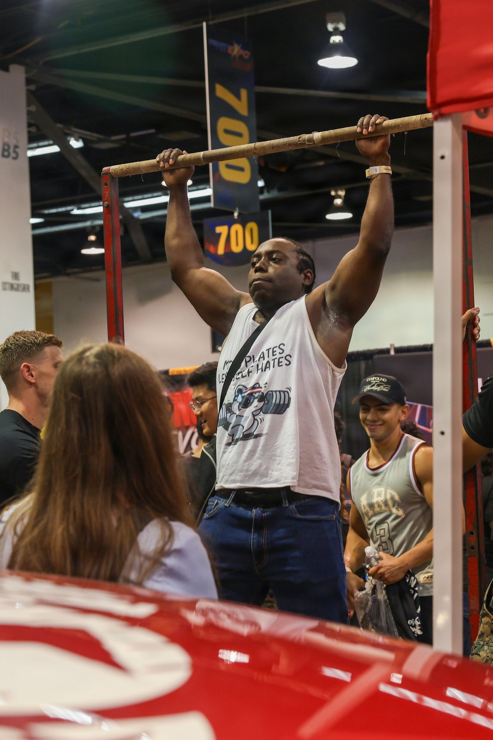 A muscular man is performing a dead hang or pull-up on a bar at what appears to be a fitness or sports expo. He is wearing a white sleeveless shirt with the text 'More Plates Less Self Hates' and is surrounded by a crowd of spectators. The setting includes banners displaying numbers like '700' and '70', suggesting a weightlifting or strength competition context.