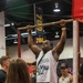 A muscular man is performing a dead hang or pull-up on a bar at what appears to be a fitness or sports expo. He is wearing a white sleeveless shirt with the text 'More Plates Less Self Hates' and is surrounded by a crowd of spectators. The setting includes banners displaying numbers like '700' and '70', suggesting a weightlifting or strength competition context.