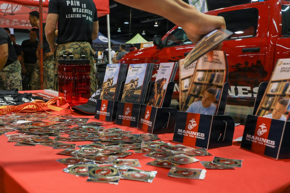 A U.S. Marines recruiting booth at what appears to be a public expo or convention. The table is covered with Marines branded materials including brochures, pamphlets, and small packaged items, likely stickers or coins. Personnel in military camouflage uniforms are visible in the background near a red vehicle.