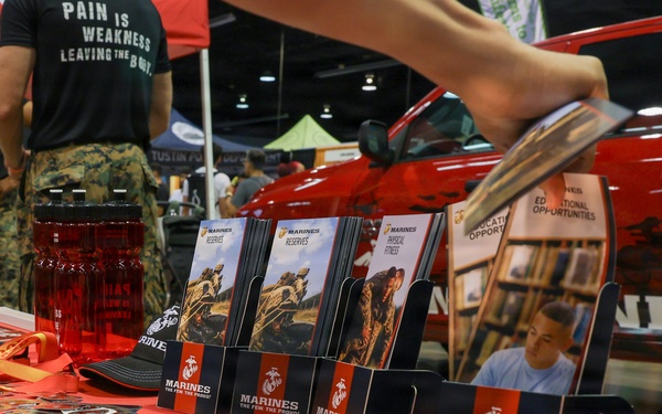 A U.S. Marines recruiting booth at what appears to be a public expo or convention. The table is covered with Marines branded materials including brochures, pamphlets, and small packaged items, likely stickers or coins. Personnel in military camouflage uniforms are visible in the background near a red vehicle.