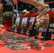 A U.S. Marines recruiting booth at what appears to be a public expo or convention. The table is covered with Marines branded materials including brochures, pamphlets, and small packaged items, likely stickers or coins. Personnel in military camouflage uniforms are visible in the background near a red vehicle.