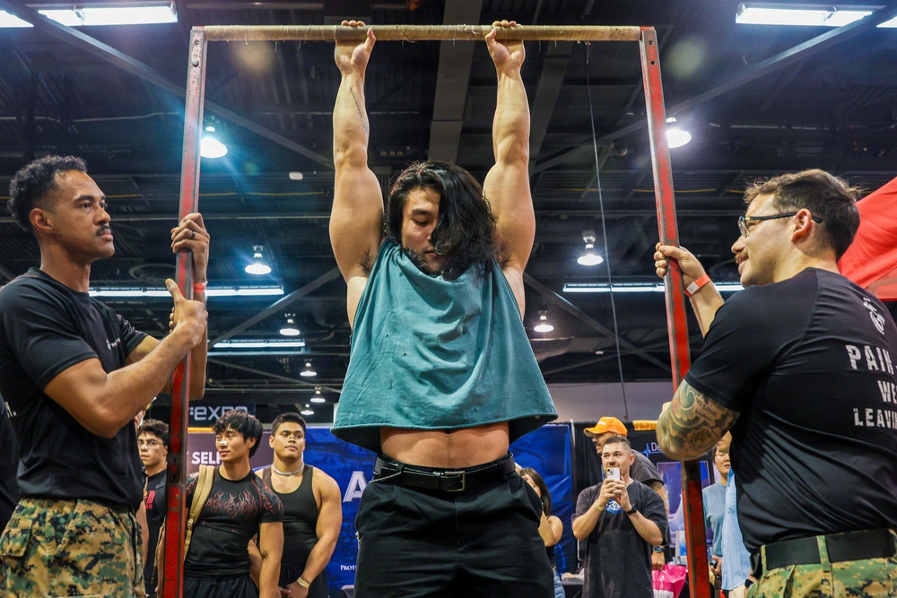 A muscular individual with long hair is performing a dead hang or pull-up on a red metal bar frame at what appears to be a fitness or military expo. Two men in black t-shirts and camouflage pants, likely military personnel, are stabilizing the frame on either side. A crowd of spectators watches in the background of what appears to be a large convention hall.