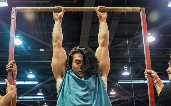 A muscular individual with long hair is performing a dead hang or pull-up on a red metal bar frame at what appears to be a fitness or military expo. Two men in black t-shirts and camouflage pants, likely military personnel, are stabilizing the frame on either side. A crowd of spectators watches in the background of what appears to be a large convention hall.