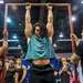 A muscular individual with long hair is performing a dead hang or pull-up on a red metal bar frame at what appears to be a fitness or military expo. Two men in black t-shirts and camouflage pants, likely military personnel, are stabilizing the frame on either side. A crowd of spectators watches in the background of what appears to be a large convention hall.