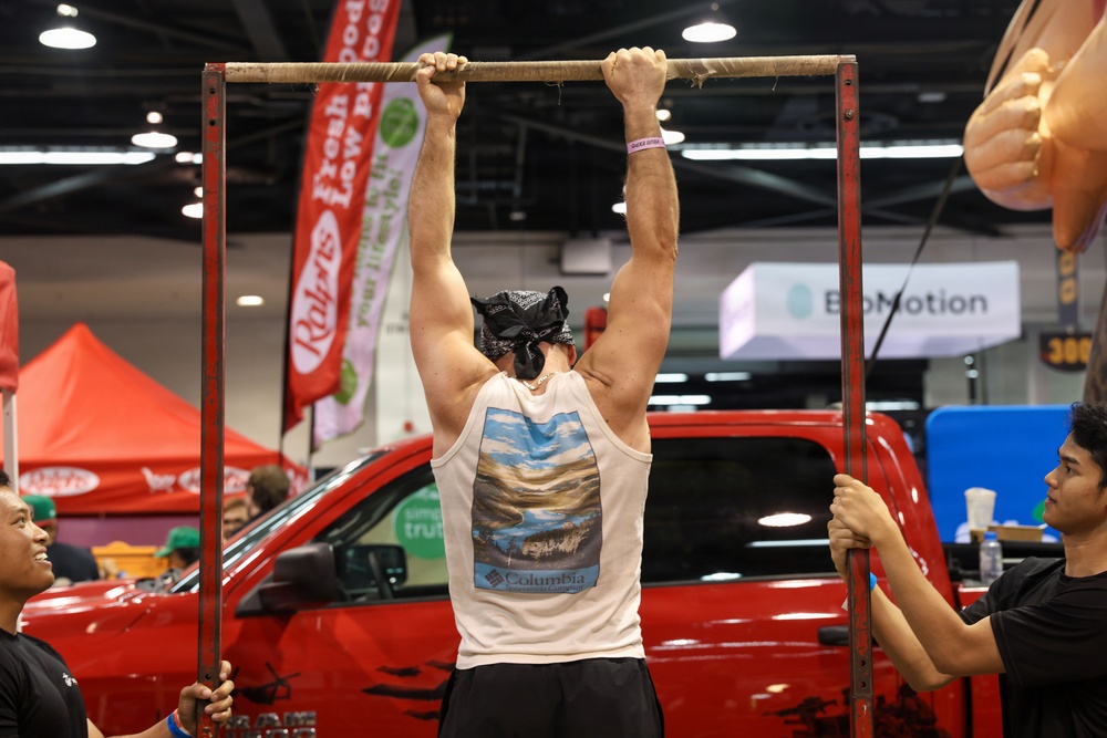 A muscular man wearing a Columbia tank top and black bandana is performing pull-ups on a makeshift bar at what appears to be an indoor expo or trade show. Two assistants are holding the metal frame steady on either side. The background features vendor booths, banners including 'Raley's' and 'BioMotion', and a red pickup truck on display.